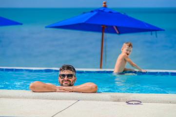 Seven years old cute boy smiling without tooth. He and his bearded father are in sunglasses, show their tongues, they leaned on pool railing giving a grimace of happiness. Sea view and beach umbrellas