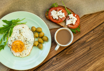 Fried eggs, a sandwich with vegetables and feta cheese and cappuccino coffee for breakfast. Tomatoes and bread on a wooden table