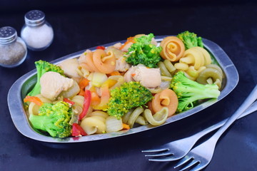 Stir fried chicken fillet with vegetables and pasta on a metal tray on a black abstract background.