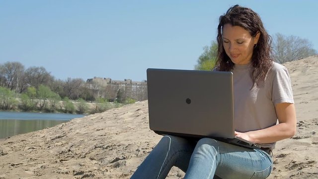 Girl with a laptop by the water. Work on the computer in the fresh air by the river. Spring sunny day in nature. Woman with a laptop on a siskin by the river.