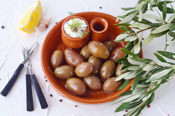 Olives in a traditional brown ceramic bowl, with tzatziki sauce, lemon, garlic, olive branch and olive oil on a white abstract background.
