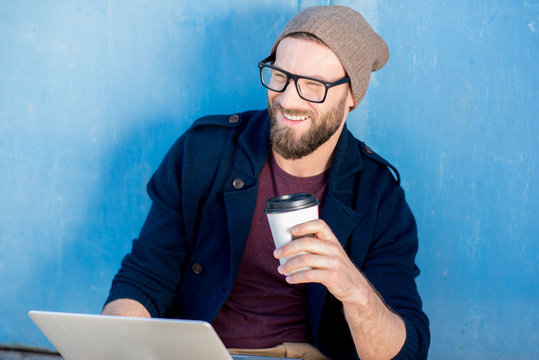Stylish man dressed casual in sweater and hat working with laptop sitting with coffee near the blue wall background