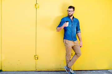 Portrait of a handsome man in blue t-shirt standing with coffee to go on the yellow background