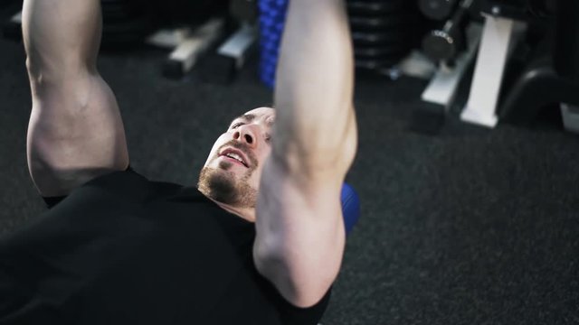 Pan shot of a bearded young man in a black T-shirt lying on a bench in a gym and doing a barbell exercise. Right to left pan real time medium shot