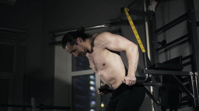 Bearded young man with a bare torso is doing push ups on parallel bars in a gym. Real time right to left pan medium shot.