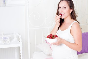 Obraz na płótnie Canvas Happy pregnant woman sitting on the bed. A young pregnant woman is sitting in the bedroom. Pregnant woman relaxing. Young pregnant woman eating strawberries.