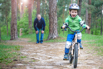 Naklejka premium Little kid boy of 3 years and his father in autumn forest with a bicycle. Dad teaching his son. Man happy about success. Child with helmet. Safety, sports, leisure with kids concept.