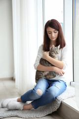 Beautiful young woman with cute cat sitting on window sill at home