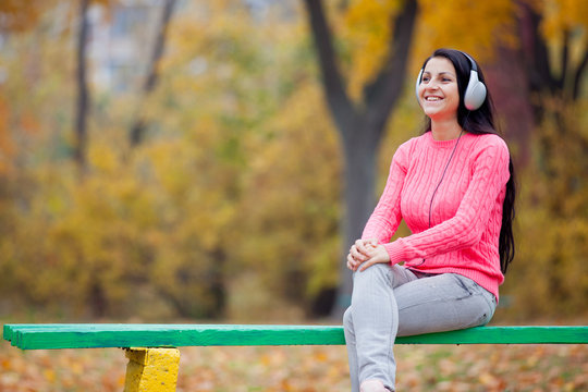 photo of beautiful young woman sitting on the bench in headphones on the wonderful yellow trees background