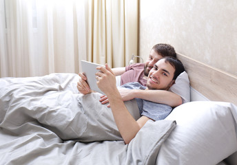 Gay couple lying on bed and using tablet at home