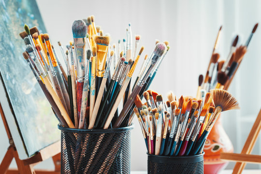 Artistic equipment in studio: wooden easel,  paintbrushes, tubes of paint, palette and paintings on work artist table.