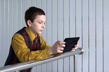 A schoolboy in a tartan waistcoat and yellow mustard shirt is reading on a tablet at the metal stair rails on the light background