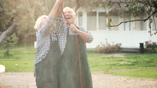 Old couple with garden hose. Cheerful woman and man outdoors.
