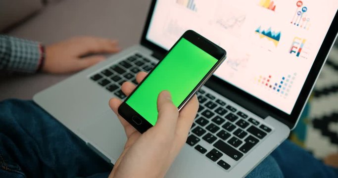Close up man hands holding smart phone with green screen. Chroma Key. Close up male hands typing on cell phone screen with laptop background with charts . Business graphics