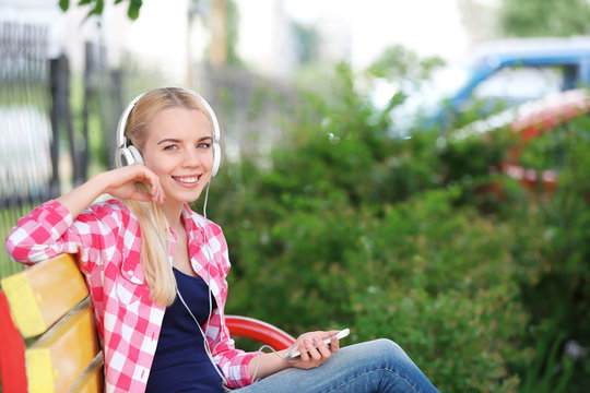 Young beautiful woman with headphones sitting on the bench and listening to music outdoors