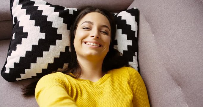 Portrait of beautiful young woman in yellow sweater lying in the pillow on the couch in the living room. Portrait of attractive woman smiling to the camera. Close up shot