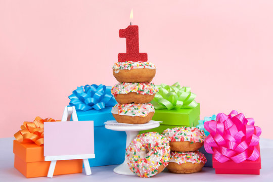 Cake donuts with white frosting and colorful sprinkles stacked on and around a small pedestal surrounded by birthday presents. Number 1 candle burning for first birthday party. Small pink easel blank