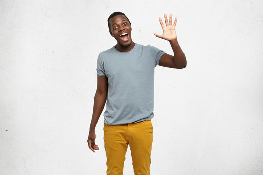 Positive human emotions, facial expressions, feelings, attitude and reaction. Friendly-looking polite young African American man dressed in grey t-shirt and mustard jeans saying hi, waving his hand
