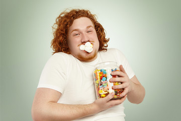 Portrait of cheerful young European overweight obese male with curly ginger hair consuming junk good, stuffing marshmallow into mouth and holding tight large glass jar, full of colorful sweets