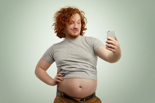 Funny red-haired overweight male trying to look attractive and sexy, holding hand on his waist while taking selfie with electronic device, belt on his pants in undone because of fat belly sticking out