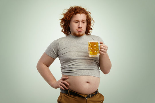 People, unhealthy lifestyle, obesity and gluttony. Overweight fat young European man with curly red hair holding glass of beer, feeling hesitated deciding whether to drink it or not after good dinner