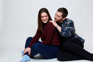 Fototapeta na wymiar A happy couple relaxing on white floor. A handsome guy wearing checked shirt touching his pretty girlfriend dressed in jeans and red sweater having smile being happy to be near her lover