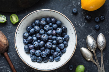Blueberries and vintage spoons: Top view of fresh blueberries in white ceramic bowl with baby kiwi fruit, lemon and vintage tea spoons on dark stone table. Healthy vegan fresh food concept
