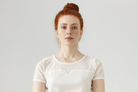Indoor portrait of attractive young European ginger woman with freckled face and hair bun standing at grey studio wall, dressed in white blouse, her look and posture expressing self-confidence