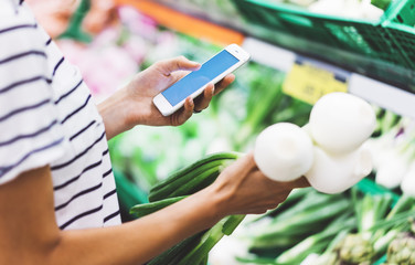 Young woman shopping healthy food in supermarket blur background. Female hands buy onion products and using smart phone in store. Hipster at grocery holding basket. Person comparing price of produce