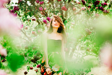 Portrait of an attractive beautiful young model girl in colorful dress with long hair in summer lilac garden with bushes and flowers posing for camera. Spring concept. Photo through the brunches.