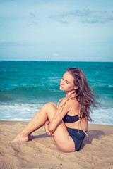 Sexy woman with tan skin in a black swimsuit sitting on the sand of the beach. Female hug her knees near sea.