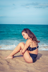 Fototapeta na wymiar Beautiful sexy woman with tan skin in a black swimsuit sitting on the sand of the beach. Female hug her knees near sea.