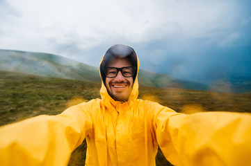 Selfie portrait of a smiling and laughing traveller man in yellow raincoat and glasses in the clouds mountains in stromy weather with rain - obrazy, fototapety, plakaty