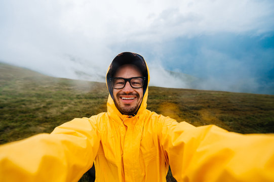 Selfie portrait of a smiling and laughing traveller man in yellow raincoat and glasses in the clouds mountains in stromy weather with rain