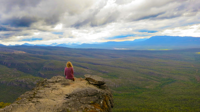 Adventurous young blonde girl sitting on a height cliff edge at the top of the Boroka Lookout at the The Grampians National Park (Gariwerd), Victoria, Australia