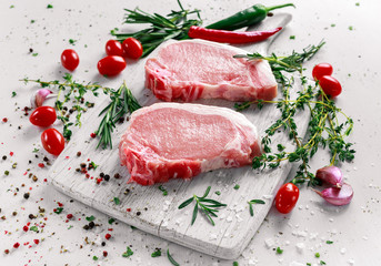 Raw Pork Loin chops on a cutting board with herbs, rosemary, thyme, chilli, salt, pepper on white cutting board.