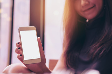 Mockup image of a beautiful woman holding and showing white mobile phone with blank white screen and smiley face in modern cafe
