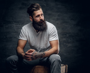 Bearded male with tattoo on arm dressed in a white t shirt sits on a wooden box.