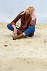 Fototapeta na wymiar Young sexy blonde girl in nature near a river and trees in American country style in a leather jacket, sweater blue jeans and brown boots with a beautiful face and pretty eyes