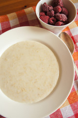 Oatmeal porridge in white round plate with red raspberry and red check tablecloth,  vertical