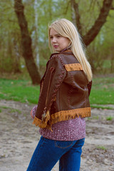 Young sexy blonde girl in nature near a river and trees in American country style in a leather jacket, sweater blue jeans and brown boots with a beautiful face and pretty eyes