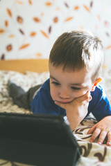 Little boy looking at tablet while lying on bed, natural light.