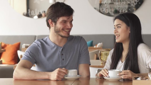 Handsome caucasian guy talking to his female friend at the cafe. Pretty asian girl smiling for the brunette bearded guy. Attractive young people holding cups of coffee in their hands