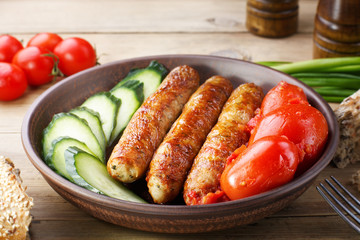 A hearty meal of roasted sausages. Fried homemade sausages with fresh cucumber and canned tomatoes in a clay bowl on a wooden table. Cherry tomatoes. Green onions.