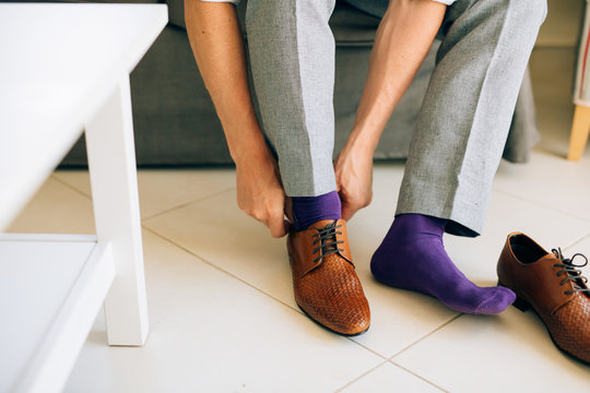 The man in gray slacks and a purple dress socks brown shoes with laces sitting on the couch.