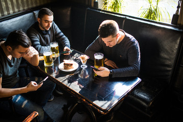 Male friends sitting in smartphones drinking beer at bar or pub. Smartphones dependence.