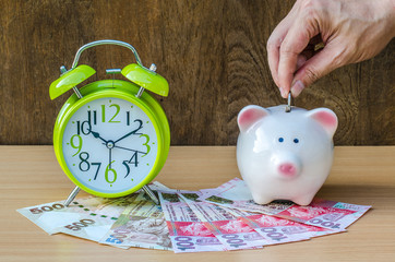 Piggy bank with money and alarm clock on wood table background ,hand add coin to piggy bank save...
