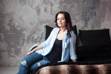 Sensual woman with dark hair in blue shirt and jeans sitting on a bed at home. Loft style interior