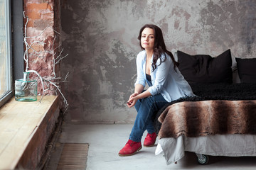 Sensual woman with dark hair in blue shirt and jeans sitting on a bed at home. Loft style interior