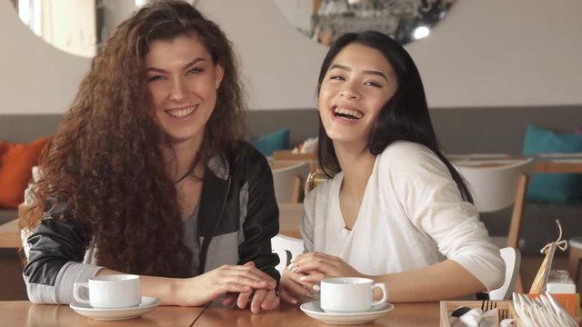 Pretty caucasian girl telling something funny to her friend. Young cute asian woman laugh about what her mate saying at the cafe. Two attractive ladies holding their hands on the table near the cups